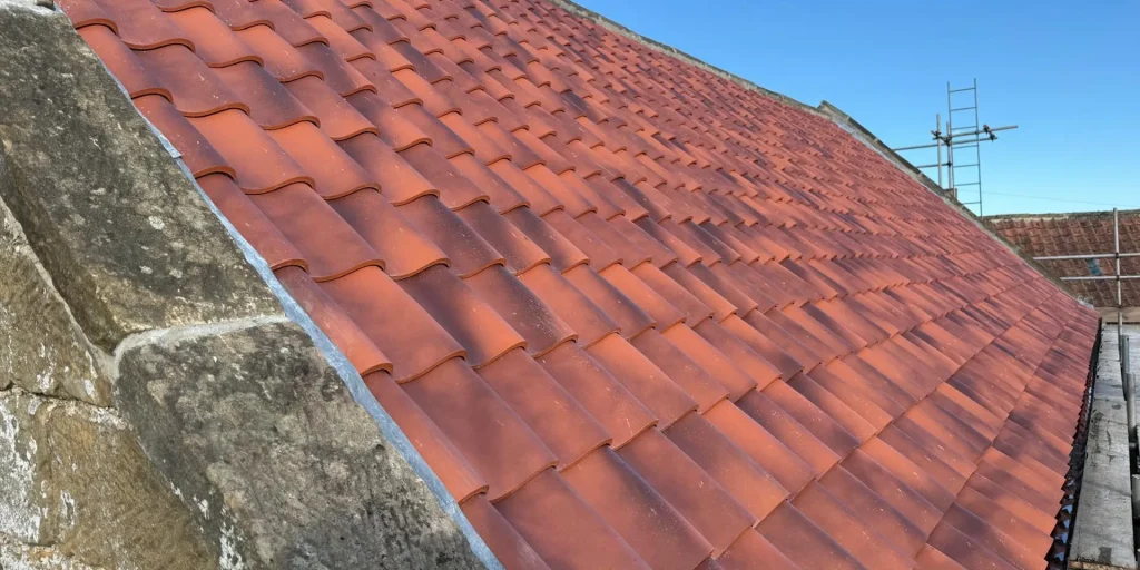 A completed red clay pantile roof with new leadwork beside a traditional stone coping, under a clear blue sky.