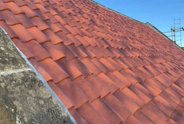 A completed red clay pantile roof with new leadwork beside a traditional stone coping, under a clear blue sky.